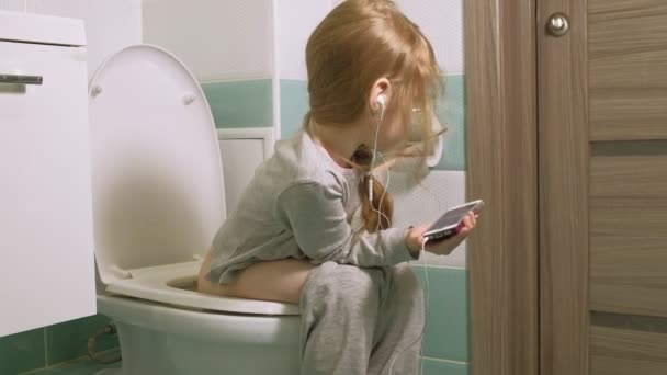 Маленькая девочка сидит на унитазе и пользуется телефоном — стоковое видео
