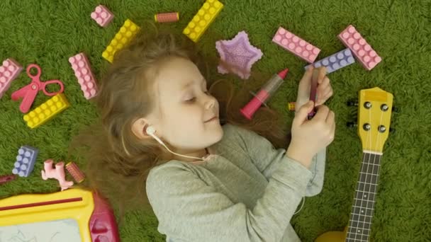 Petite fille couchée sur un fond vert dans les écouteurs et à l'aide d'un téléphone, vue de dessus — Video