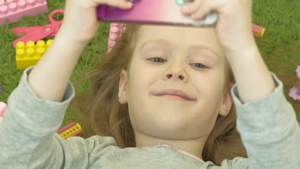 Маленька дівчинка лежить на зеленому фоні в навушниках і використовує телефон, вид зверху — стокове відео