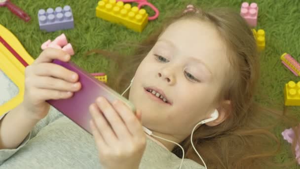 Petite fille couchée sur un fond vert dans les écouteurs et à l'aide d'un téléphone, vue de dessus — Video