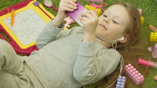 Bambina sdraiata su uno sfondo verde in cuffia e utilizzando un telefono, vista dall'alto — Video Stock