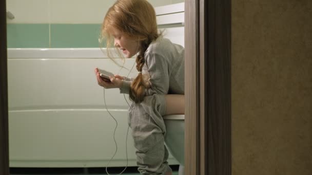 Petite fille s'assoit sur les toilettes et utilise le téléphone — Video