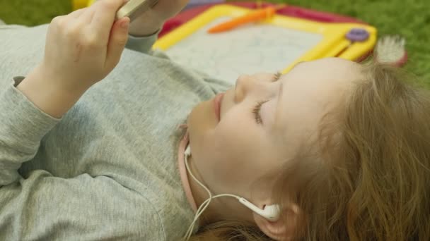 Niña acostada sobre un fondo verde en auriculares y usando un teléfono, vista superior — Vídeos de Stock