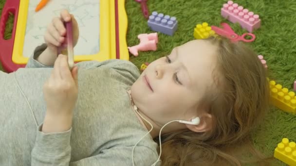 Petite fille couchée sur un fond vert dans les écouteurs et à l'aide d'un téléphone, vue de dessus — Video