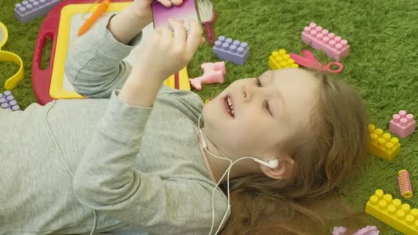 Bambina sdraiata su uno sfondo verde in cuffia e utilizzando un telefono, vista dall'alto — Video Stock
