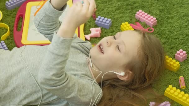 Little girl lying on a green background in headphones and using a phone, top view — Stock Video