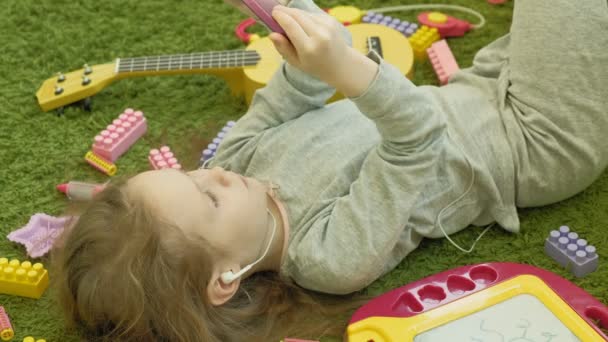 Little girl lying on a green background in headphones and using a phone, top view — Stock Video