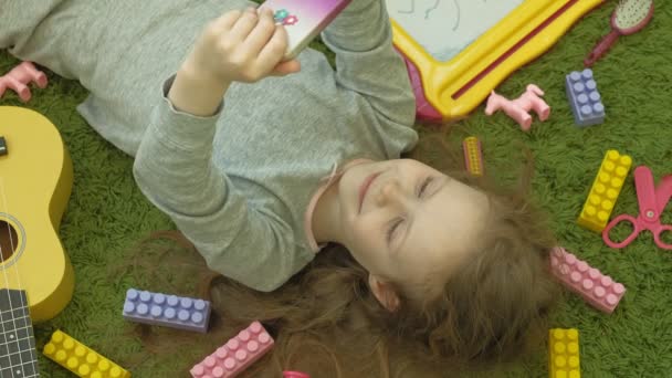 Kis lány feküdt a zöld háttér fejhallgató és telefon használata, felülnézet — Stock videók