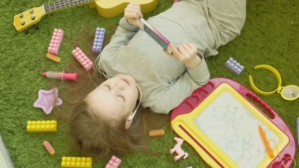 Liten flicka som ligger på en grön bakgrund i hörlurar och med hjälp av en telefon, uppifrån — Stockvideo
