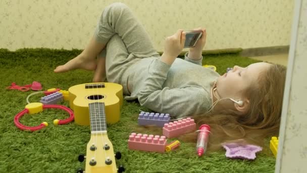 Kleines Mädchen, das mit Kopfhörern auf grünem Hintergrund liegt und ein Telefon benutzt, Draufsicht — Stockvideo