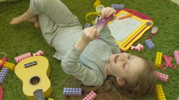 Little girl lying on a green background in headphones and using a phone, top view — Stock Video