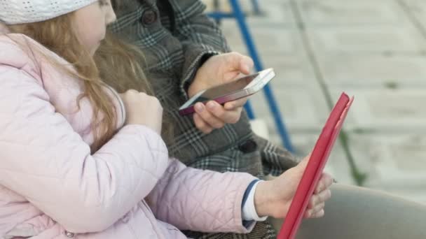 Frau mit kleinem Mädchen sitzt auf einer Bank und benutzt Gadgets — Stockvideo