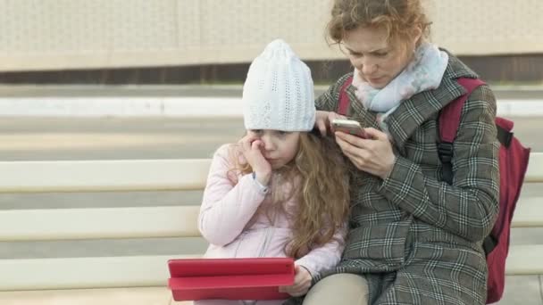 Mujer con una niña está sentado en un banco y el uso de gadgets — Vídeos de Stock