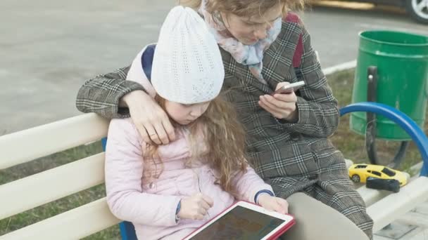 Mujer con una niña está sentado en un banco y el uso de gadgets — Vídeos de Stock