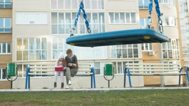 Femme avec une petite fille est assis sur un banc et en utilisant des gadgets — Video