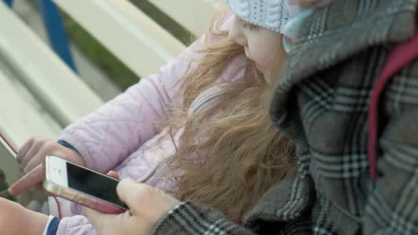 Femme avec une petite fille est assis sur un banc et en utilisant des gadgets — Video