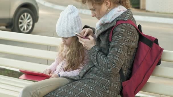 Kobieta z małą dziewczynką siedzi na ławce i za pomocą gadżety — Wideo stockowe