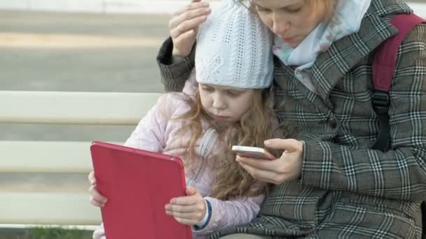 Femme avec une petite fille est assis sur un banc et en utilisant des gadgets — Video