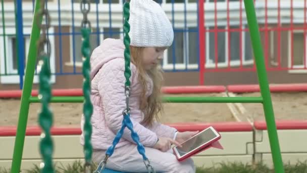 Petite fille assise sur une balançoire sur l'aire de jeux près de l'immeuble, utilise une tablette d'ordinateur — Video