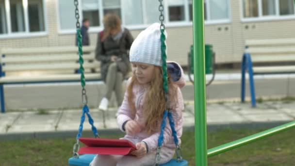 Little girl sitting on a swing on the playground near the apartment building, uses a computer tablet — Stock Video