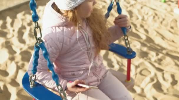 Niña montando un columpio y usando un teléfono con auriculares — Vídeo de stock