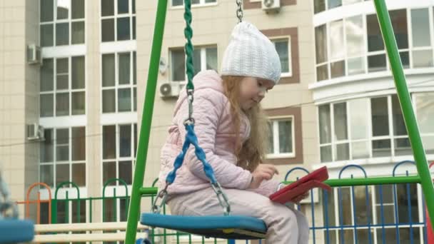 Menina sentada em um balanço no playground perto do prédio do apartamento, usa um tablet de computador — Vídeo de Stock