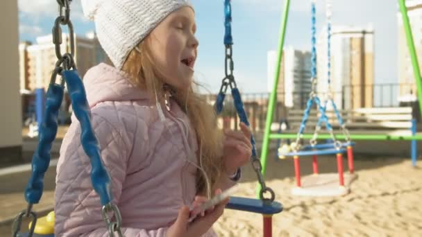 Petite fille chevauchant une balançoire et utilisant un téléphone avec écouteurs — Video