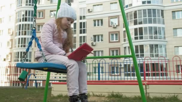 Klein meisje zittend op een schommel op de speelplaats in de buurt van het appartementencomplex, maakt gebruik van een computer Tablet — Stockvideo