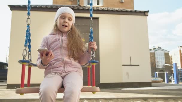 Little girl riding a swing and using a phone with headphones — Stock Video