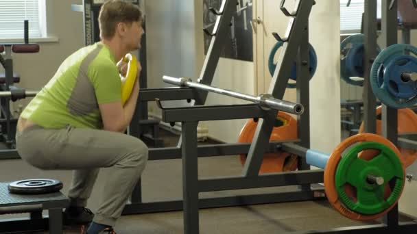 El hombre con sobrepeso hace sentadillas con sentadillas con un disco de peso para una barra. Entrenamiento físico. concepto de estilo de vida saludable — Vídeos de Stock