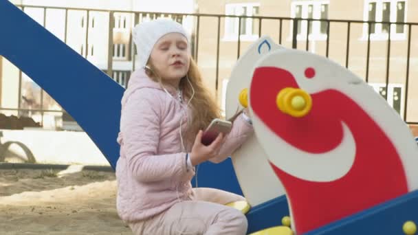 Niña montando un columpio y usando un teléfono con auriculares — Vídeo de stock