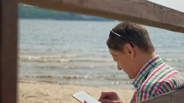 Un hombre en la playa utiliza una tableta de ordenador . — Vídeos de Stock