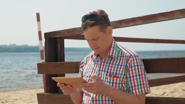 Un hombre en la playa utiliza una tableta de ordenador . — Vídeos de Stock