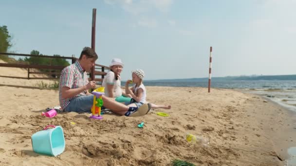 Dojrzały mężczyzna w koszuli z dwiema dziewczynami siedzi na piasku nad rzeką. Rodzinne zajęcia na świeżym powietrzu — Wideo stockowe