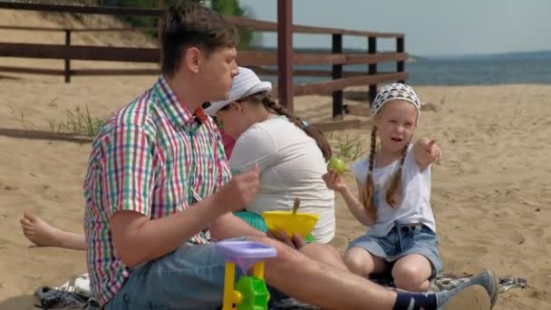 A mature man in a plaid shirt with two girls are sitting on the sand by the river. Family outdoor activities — Stock Video