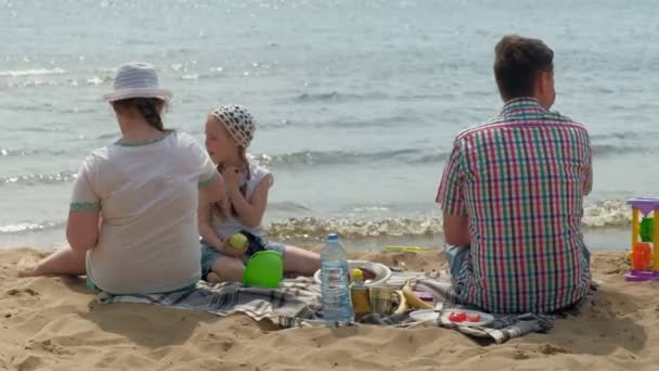 Um homem maduro com uma camisa xadrez com duas meninas está sentado na areia junto ao rio. Família atividades ao ar livre — Vídeo de Stock