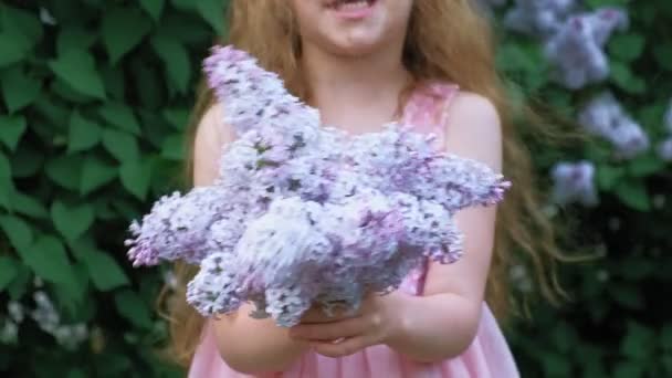 A little girl outdoors in a park or garden holds lilac flowers. Lilac bushes in the background. Summer, park — Stock Video