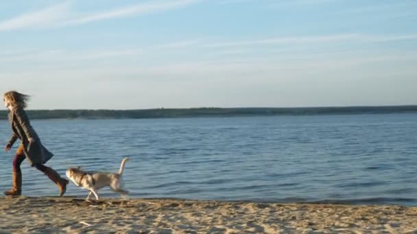 一个年轻女子在河岸的海滩上跑着，和一只棕色的狗拉布拉多玩耍。春天. — 图库视频影像