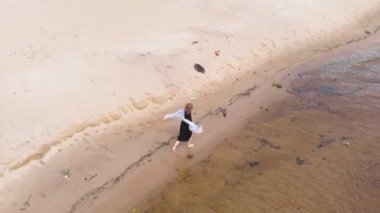 Elbiseli genç bir kadın sahil boyunca koşuyor. Hava çekimi