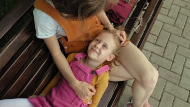 Maman avec sa fille, fille d'âge préscolaire allongée sur les genoux d'une jeune femme dans le parc sur le banc. Journée ensoleillée d'été — Video