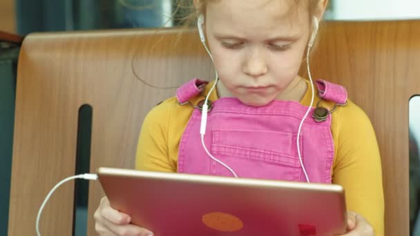 Preschool girl uses headphones and a computer tablet outdoors. light day time — Stock Video