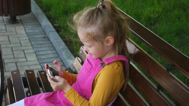Ένα χαμογελαστή κορίτσι μια προσχολική ηλικία, χρησιμοποιεί το τηλέφωνο σε εξωτερικούς χώρους στο πάρκο. Ηλιόλουστη καλοκαιρινή μέρα. — Αρχείο Βίντεο