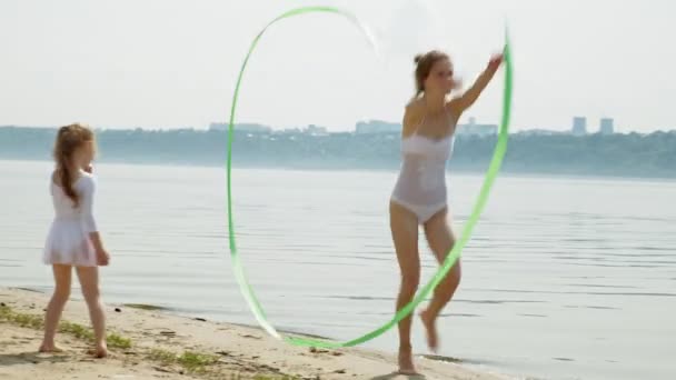 Moeder en dochter in witte badpakken dansen met gymnastisch lint op een zandstrand. Zomer, Dawn — Stockvideo