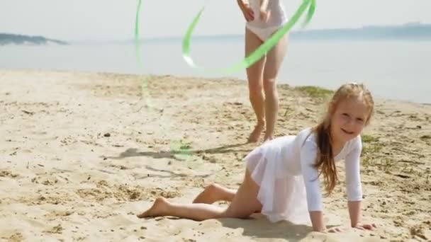 Мать и дочь в белых купальниках танцуют с гимнастической лентой на песчаном пляже. Лето, рассвет — стоковое видео