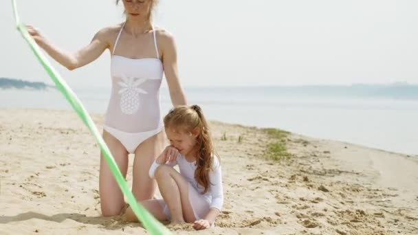 Ibu dan anak dalam pakaian renang putih menari dengan pita senam di pantai berpasir. Musim panas, fajar — Stok Video