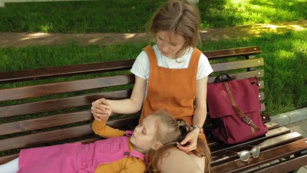 Мама с дочерью, дошкольница лежит на коленях у молодой женщины в парке на скамейке запасных. Солнечный летний день — стоковое видео