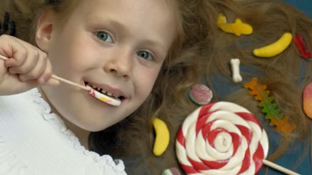 Little girl with a lollipop lies on a blue background. Closeup portrait, top view — Stock Video