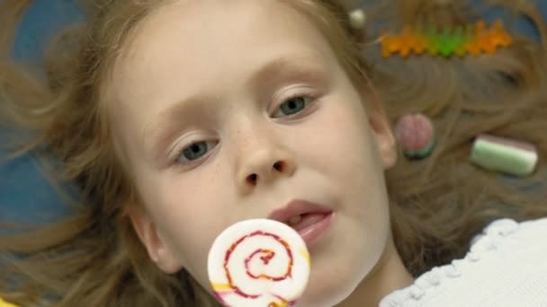 La niña con una piruleta yace sobre un fondo azul. Retrato de primer plano, vista superior — Vídeos de Stock