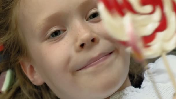 La niña con una piruleta yace sobre un fondo azul. Retrato de primer plano, vista superior — Vídeos de Stock