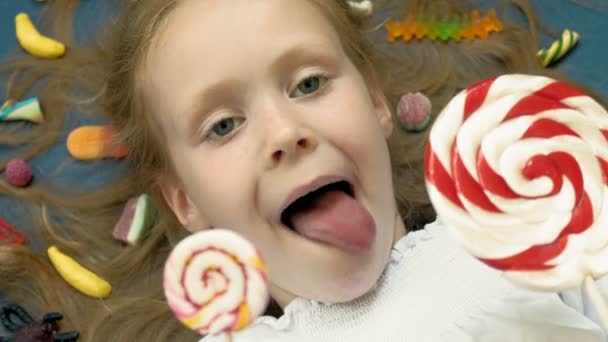 Little girl with a lollipop lies on a blue background. Closeup portrait, top view — Stock Video